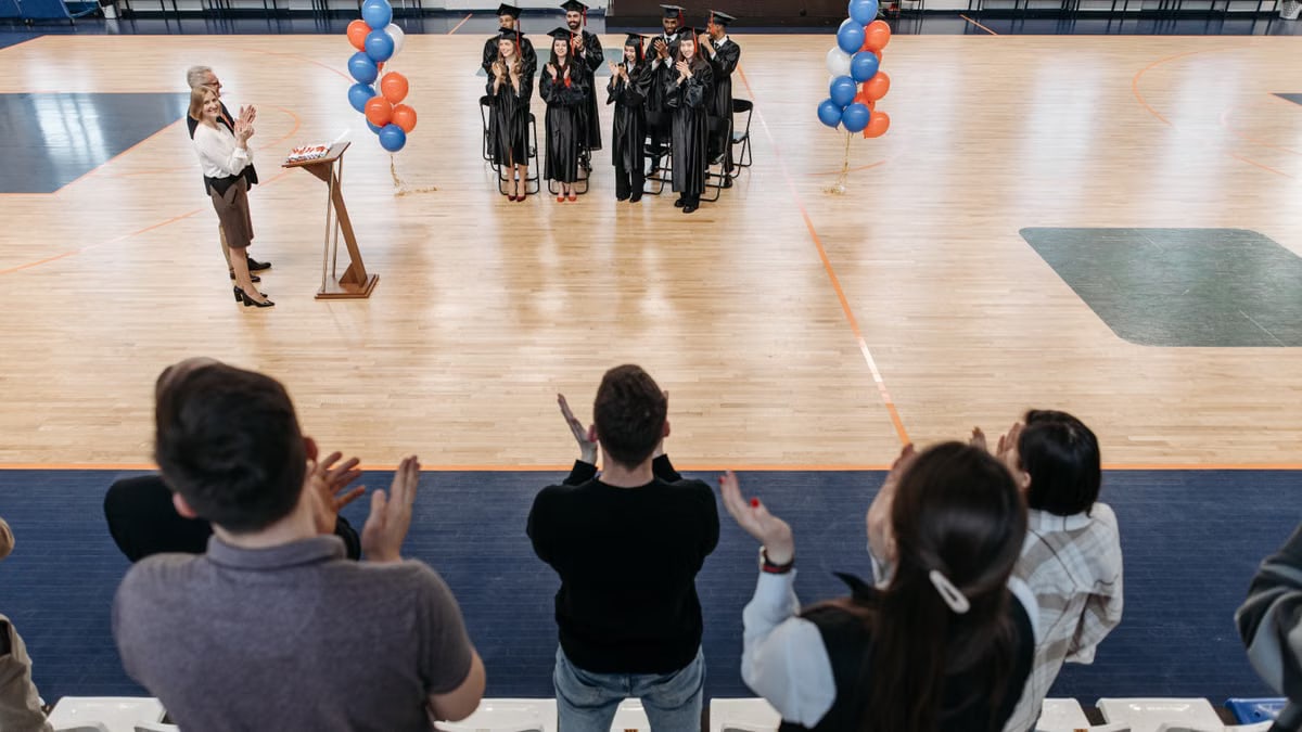 Graduates standing and clapping in a gym after receiving diplomas during a graduation ceremony.